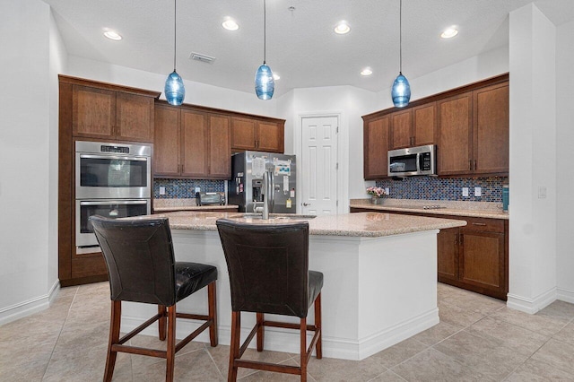 kitchen featuring decorative backsplash, hanging light fixtures, appliances with stainless steel finishes, an island with sink, and light stone counters