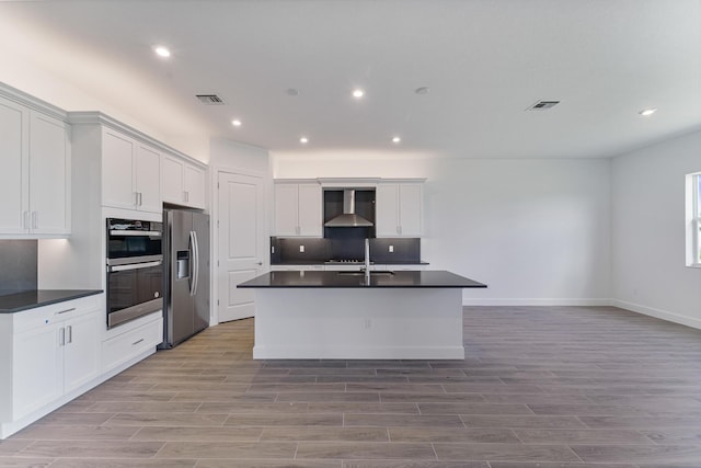 kitchen with white cabinetry, appliances with stainless steel finishes, wall chimney range hood, and an island with sink