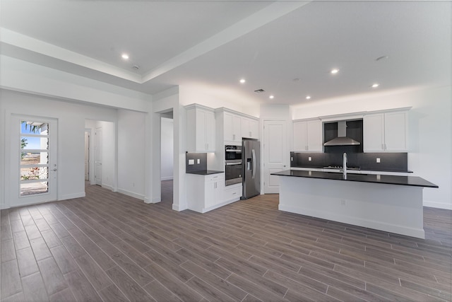 kitchen featuring wall chimney exhaust hood, white cabinetry, a center island with sink, stainless steel appliances, and decorative backsplash