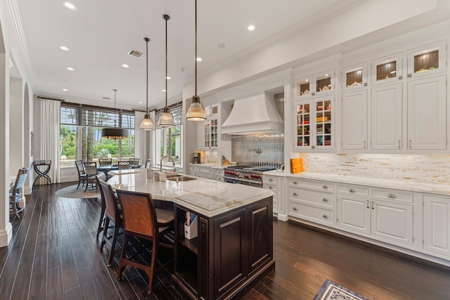 kitchen featuring backsplash, a kitchen island with sink, custom exhaust hood, white cabinetry, and sink