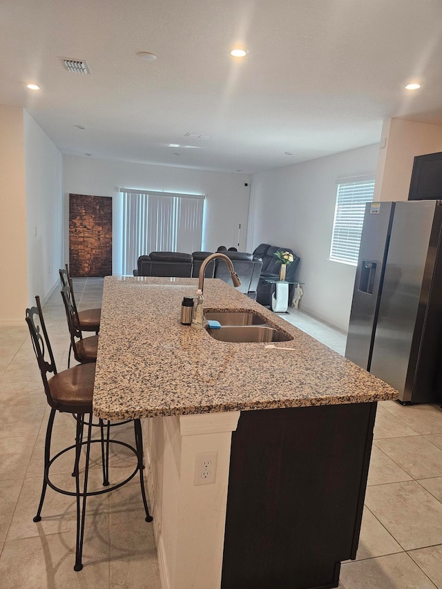 kitchen with a center island with sink, stainless steel refrigerator with ice dispenser, light stone counters, sink, and a kitchen bar