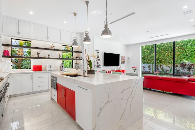 kitchen with an island with sink, decorative light fixtures, light tile flooring, light stone counters, and white cabinets