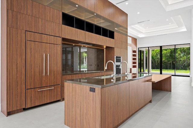 kitchen featuring a high ceiling, an island with sink, a tray ceiling, double oven, and sink