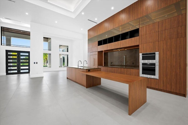 kitchen with a high ceiling, an island with sink, wood walls, and double oven