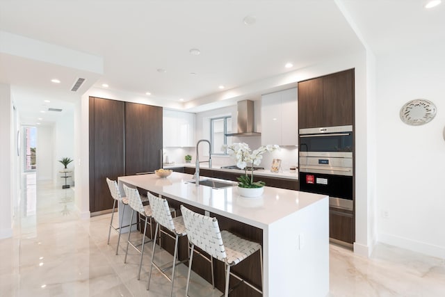 kitchen featuring a kitchen breakfast bar, an island with sink, white cabinetry, and wall chimney range hood