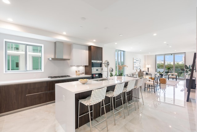 kitchen with wall chimney exhaust hood, gas cooktop, a kitchen bar, a center island with sink, and white cabinets