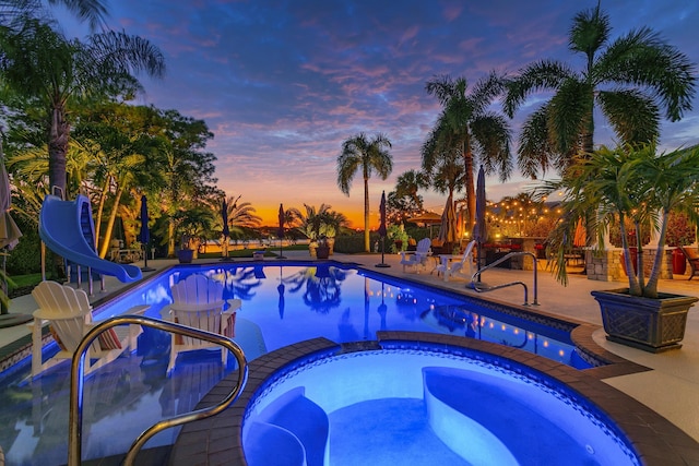 pool at dusk with a water slide, an in ground hot tub, and a patio