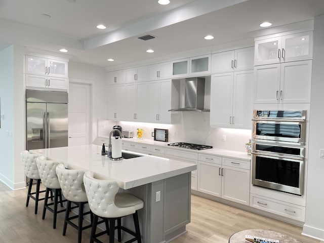 kitchen featuring wall chimney exhaust hood, stainless steel appliances, sink, white cabinets, and an island with sink