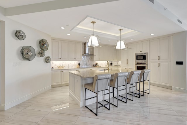 kitchen with light stone countertops, stainless steel appliances, wall chimney range hood, an island with sink, and a tray ceiling