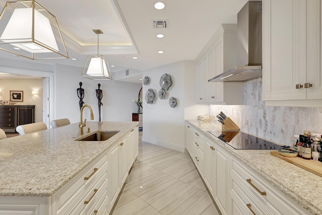 kitchen with black electric cooktop, sink, wall chimney range hood, hanging light fixtures, and an island with sink