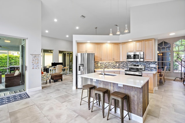 kitchen with appliances with stainless steel finishes, tasteful backsplash, a kitchen island with sink, light brown cabinets, and pendant lighting