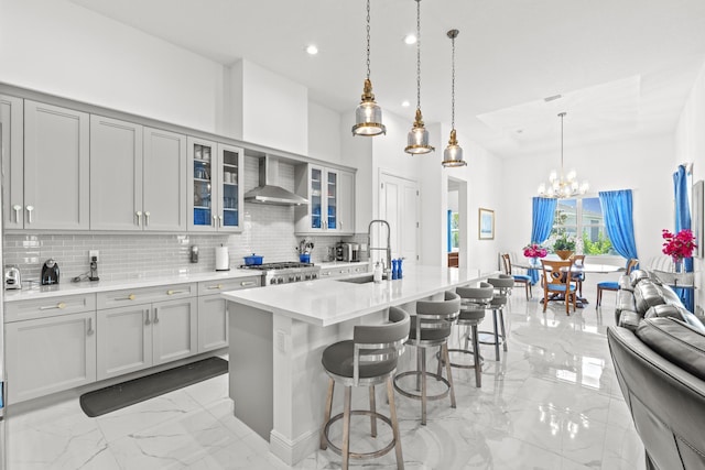 kitchen featuring wall chimney range hood, gray cabinets, an island with sink, tasteful backsplash, and a kitchen bar