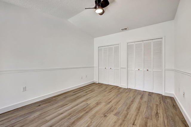 unfurnished bedroom with ceiling fan, light hardwood / wood-style floors, a textured ceiling, vaulted ceiling, and two closets