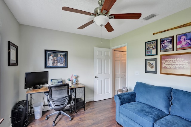 home office with a textured ceiling, a ceiling fan, visible vents, baseboards, and dark wood finished floors