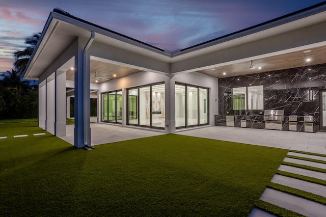 back house at dusk featuring a lawn, an outdoor kitchen, ceiling fan, and a patio area