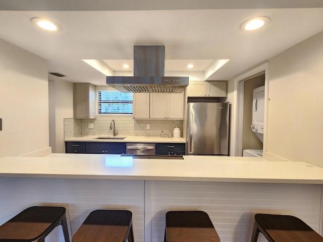 kitchen featuring sink, stacked washing maching and dryer, stainless steel appliances, island exhaust hood, and a breakfast bar