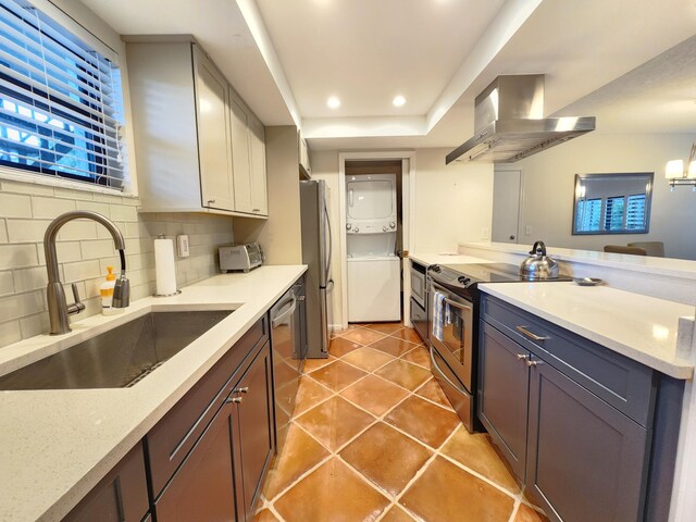 kitchen with sink, wall chimney exhaust hood, decorative backsplash, stacked washing maching and dryer, and appliances with stainless steel finishes