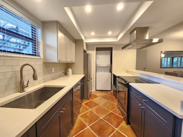 kitchen featuring backsplash, stacked washing maching and dryer, stainless steel appliances, sink, and wall chimney range hood