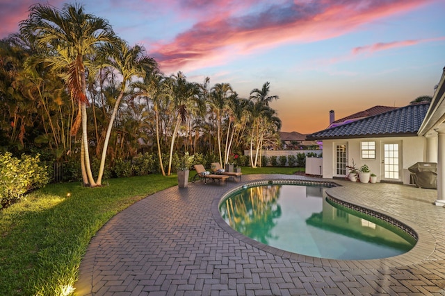 pool at dusk with french doors, a patio area, and a lawn