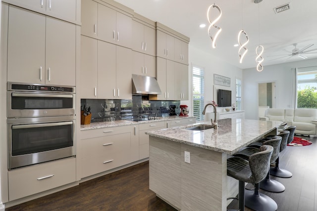 kitchen with sink, a kitchen island with sink, light stone countertops, decorative light fixtures, and stainless steel double oven