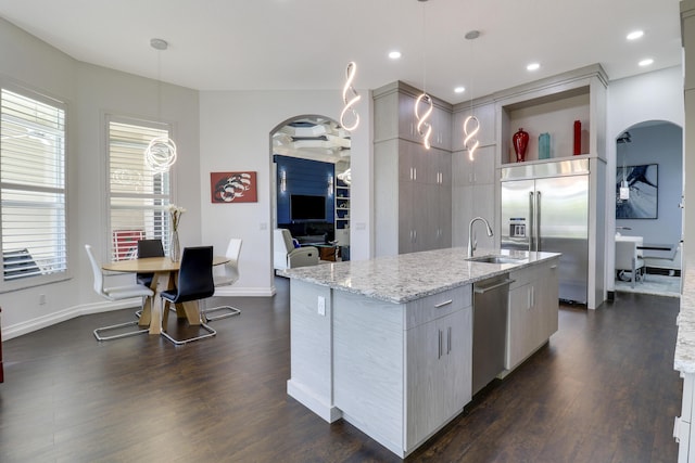 kitchen featuring dark wood-type flooring, pendant lighting, stainless steel appliances, light stone countertops, and a kitchen island with sink