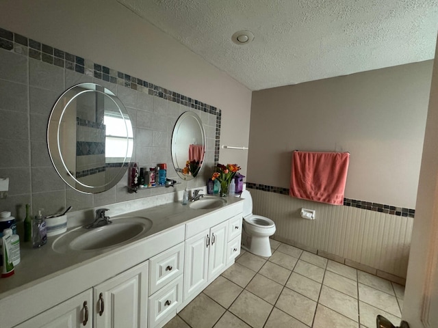 bathroom with a textured ceiling, tile floors, dual bowl vanity, tile walls, and toilet