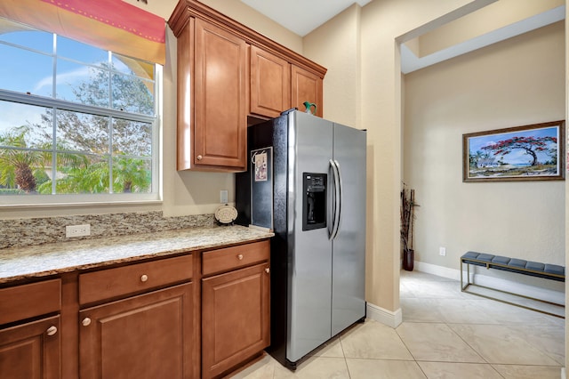 kitchen with light stone counters, brown cabinets, stainless steel refrigerator with ice dispenser, light tile patterned flooring, and baseboards