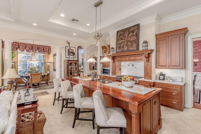 interior space featuring pendant lighting, sink, decorative columns, an island with sink, and a raised ceiling