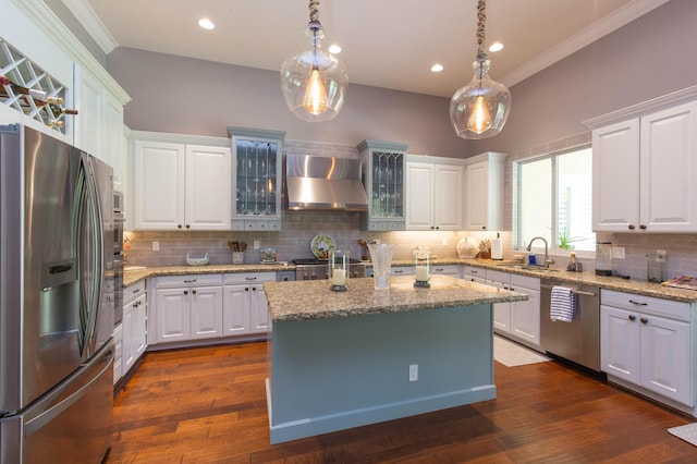 kitchen featuring an island with sink, appliances with stainless steel finishes, wall chimney range hood, and hanging light fixtures