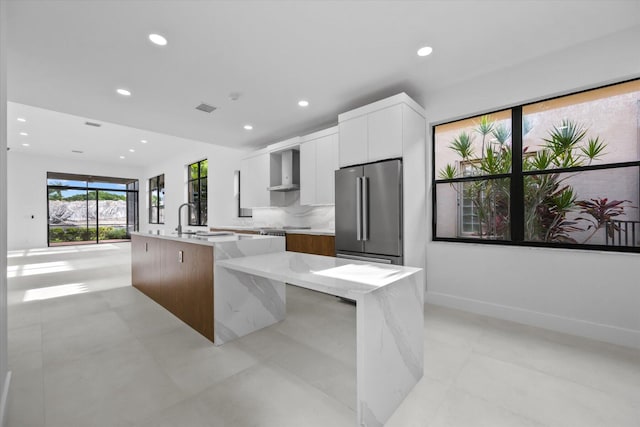 kitchen with a large island, wall chimney exhaust hood, light stone counters, stainless steel fridge, and white cabinets