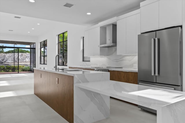 kitchen featuring light stone countertops, white cabinets, an island with sink, and wall chimney range hood