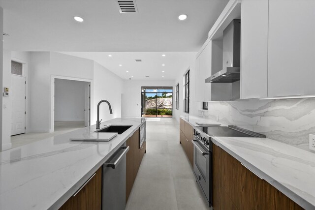 kitchen featuring light stone countertops, appliances with stainless steel finishes, sink, wall chimney range hood, and white cabinets