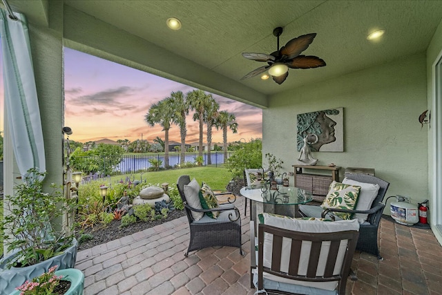 patio terrace at dusk with ceiling fan, a lawn, and a water view