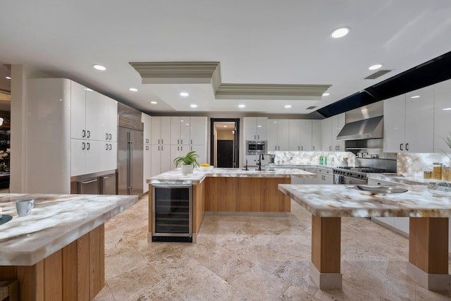 kitchen with white cabinets, wine cooler, built in appliances, wall chimney exhaust hood, and an island with sink