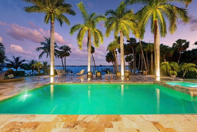 pool at dusk featuring an in ground hot tub, a water view, and a patio area