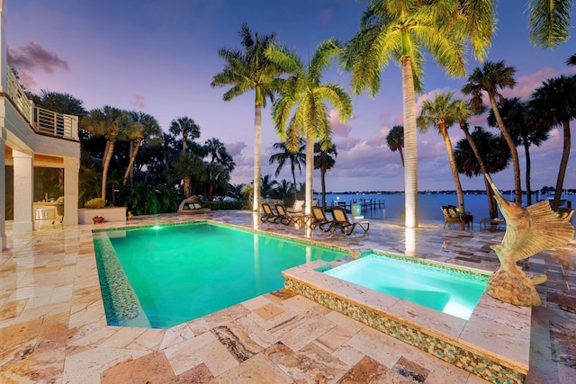 pool at dusk with an in ground hot tub, a patio, and a water view