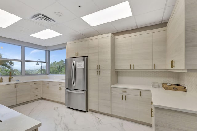 kitchen featuring light brown cabinetry, sink, a drop ceiling, and stainless steel refrigerator