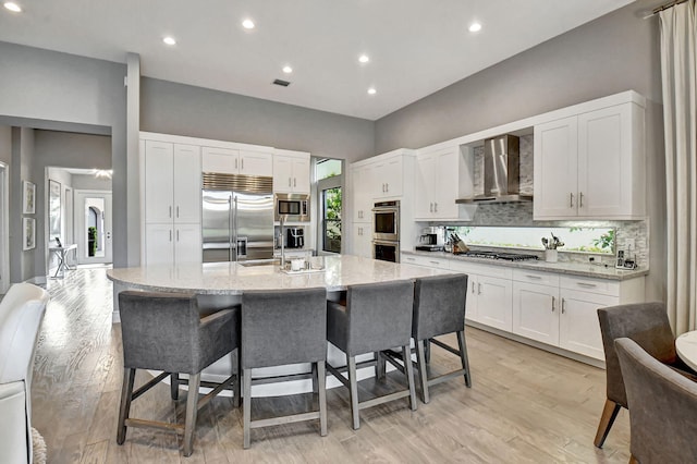 kitchen with wall chimney range hood, backsplash, built in appliances, an island with sink, and light wood-type flooring