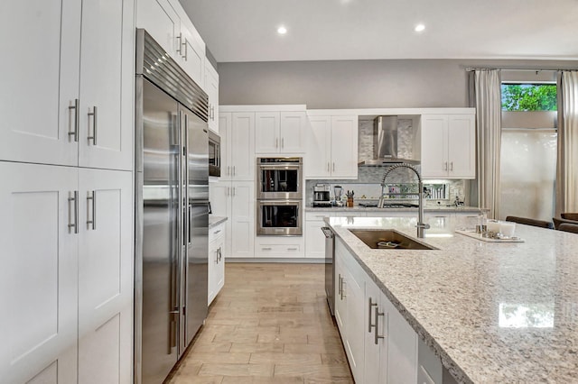 kitchen with light stone countertops, built in appliances, backsplash, wall chimney exhaust hood, and white cabinetry