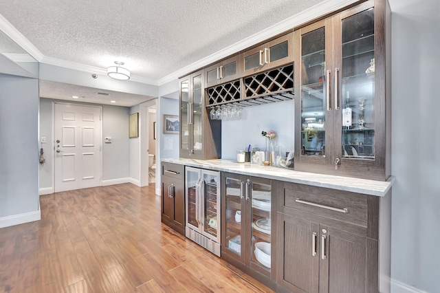 bar with ornamental molding, dark brown cabinets, light wood-type flooring, and beverage cooler