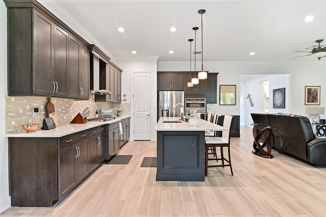 kitchen with decorative light fixtures, ornamental molding, an island with sink, stainless steel appliances, and wall chimney range hood