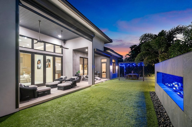 back house at dusk featuring an outdoor hangout area, a lawn, a patio, and ceiling fan