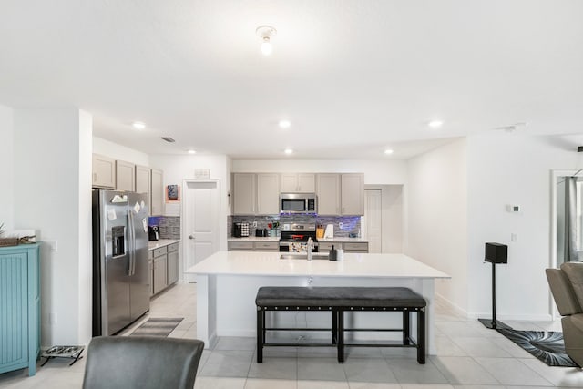 kitchen with gray cabinets, a kitchen island with sink, decorative backsplash, stainless steel appliances, and a kitchen breakfast bar