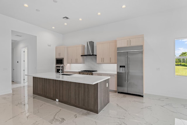 kitchen with a center island with sink, built in appliances, light countertops, wall chimney range hood, and a sink