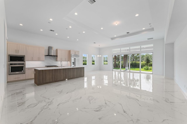 kitchen with a raised ceiling, wall chimney exhaust hood, built in appliances, a kitchen island with sink, and light countertops