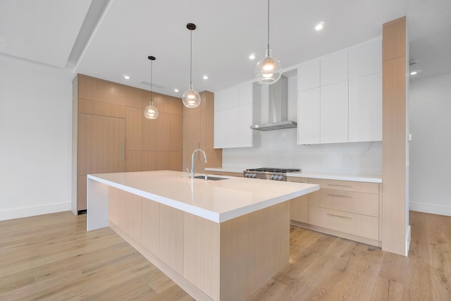 kitchen featuring an island with sink, sink, white cabinets, wall chimney range hood, and pendant lighting