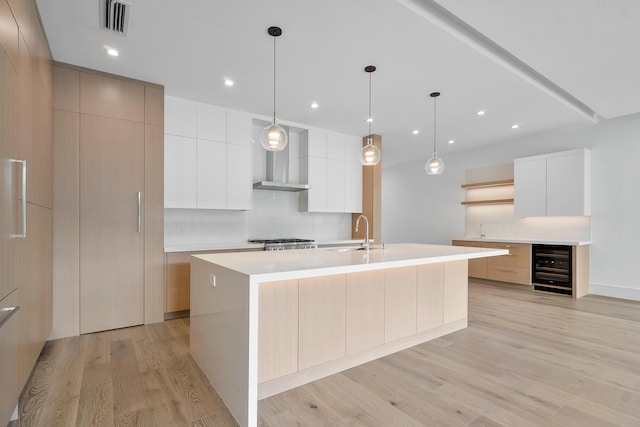 kitchen with wall chimney range hood, pendant lighting, white cabinetry, a kitchen island with sink, and wine cooler