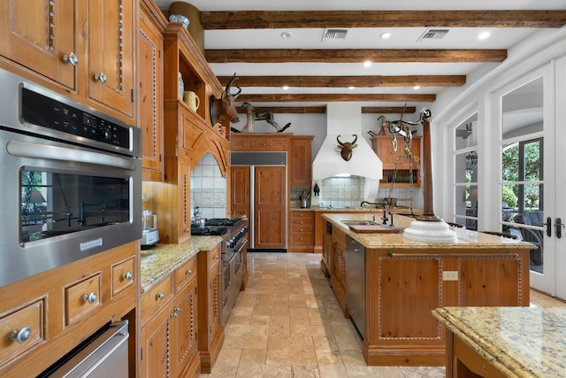 kitchen with light stone countertops, tasteful backsplash, premium range hood, a kitchen island with sink, and appliances with stainless steel finishes