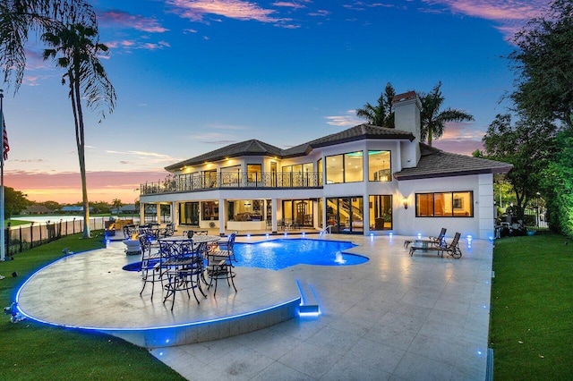 back of property at dusk featuring a patio, a balcony, fence, an outdoor pool, and a chimney