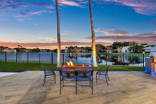 patio terrace at dusk with a water view, fence, a fire pit, and a lawn